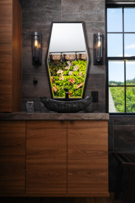 Custom wooden vanity with cabinetry, stone countertops, and a mirror with two sconces on either side. Plant wall in the reflection of the mirror. Window to the right of the image.